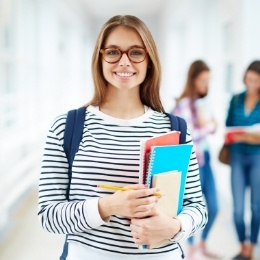 Smiling young woman wearing backpack and carrying books in school hallway